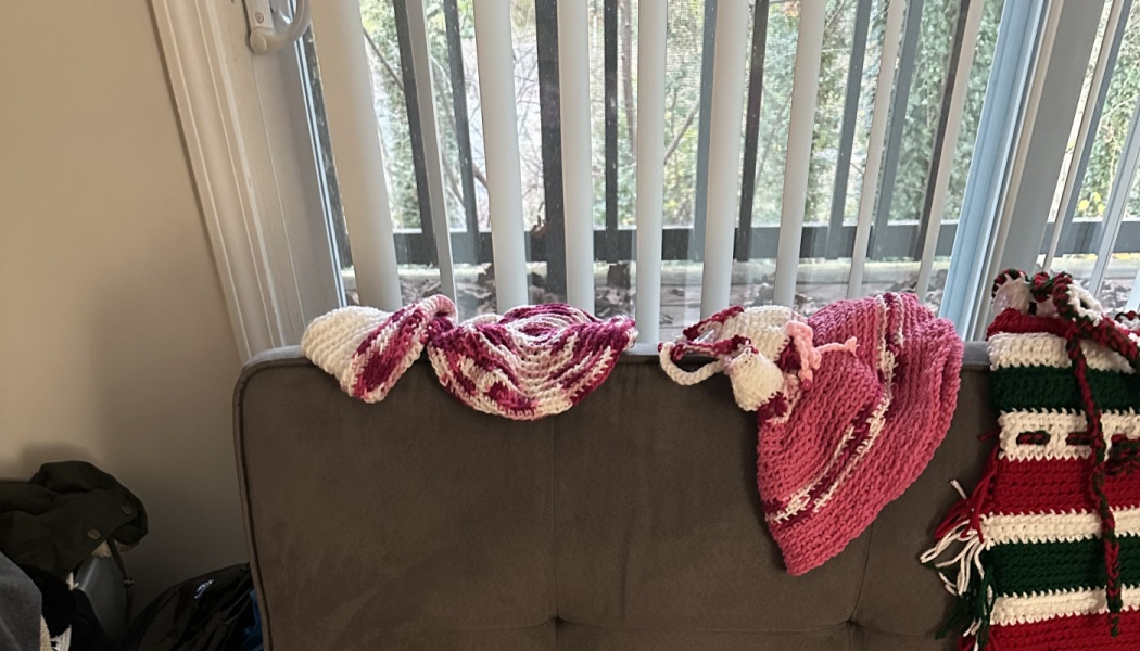 Handmade pink and white crocheted hats and a red-and-green striped stocking resting on the top of a grey couch.