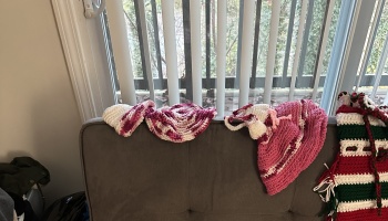Handmade pink and white crocheted hats and a red-and-green striped stocking resting on the top of a grey couch.