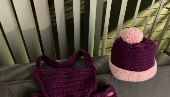 Handmade purple crocheted baby overalls alongside a matching beanie with a pink pom-pom and brim.