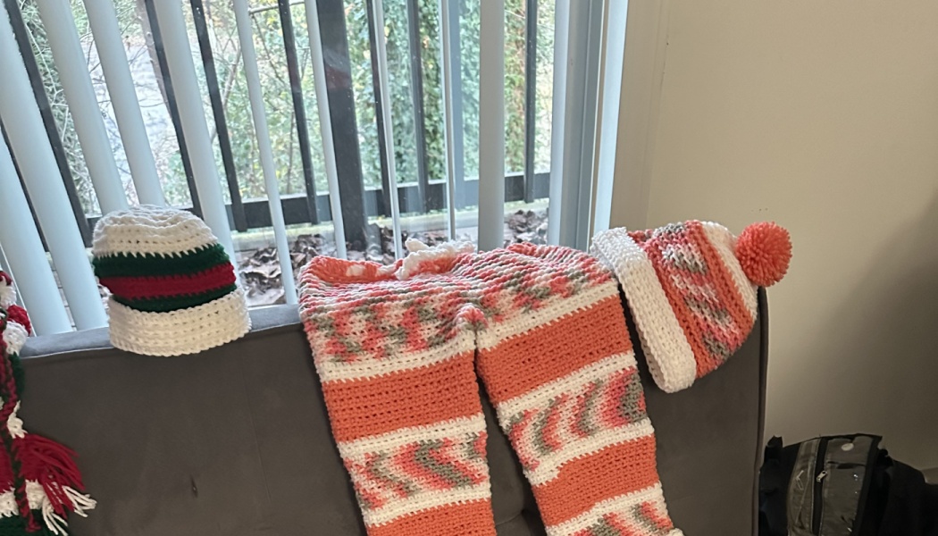 Crocheted coral and white striped pants, a matching pom-pom hat, and a white, green, and red beanie on a sofa.