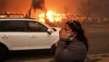 A distressed woman covers her face in tears as a raging wildfire burns homes in the background.
