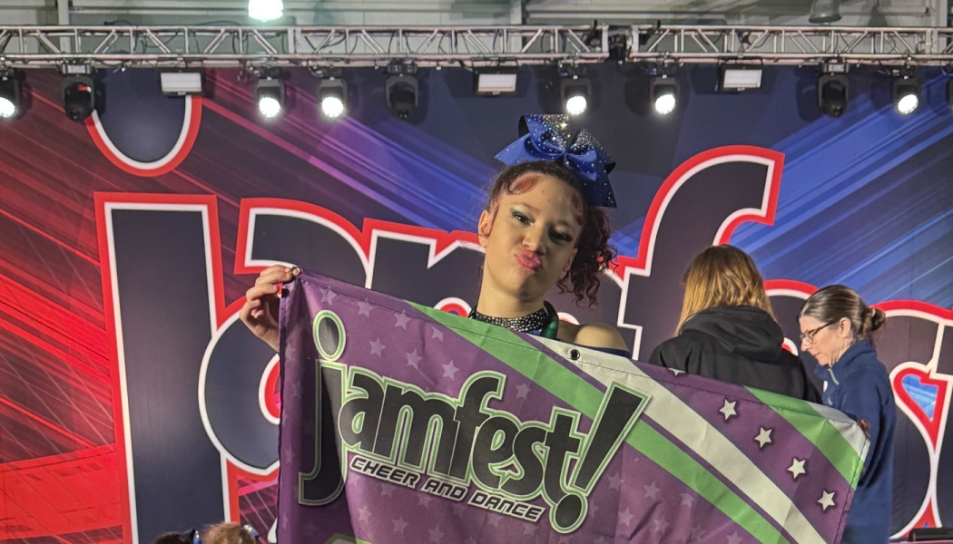 Cheerleader with a large blue bow holding a purple Jamfest Cheer and Dance banner on stage.