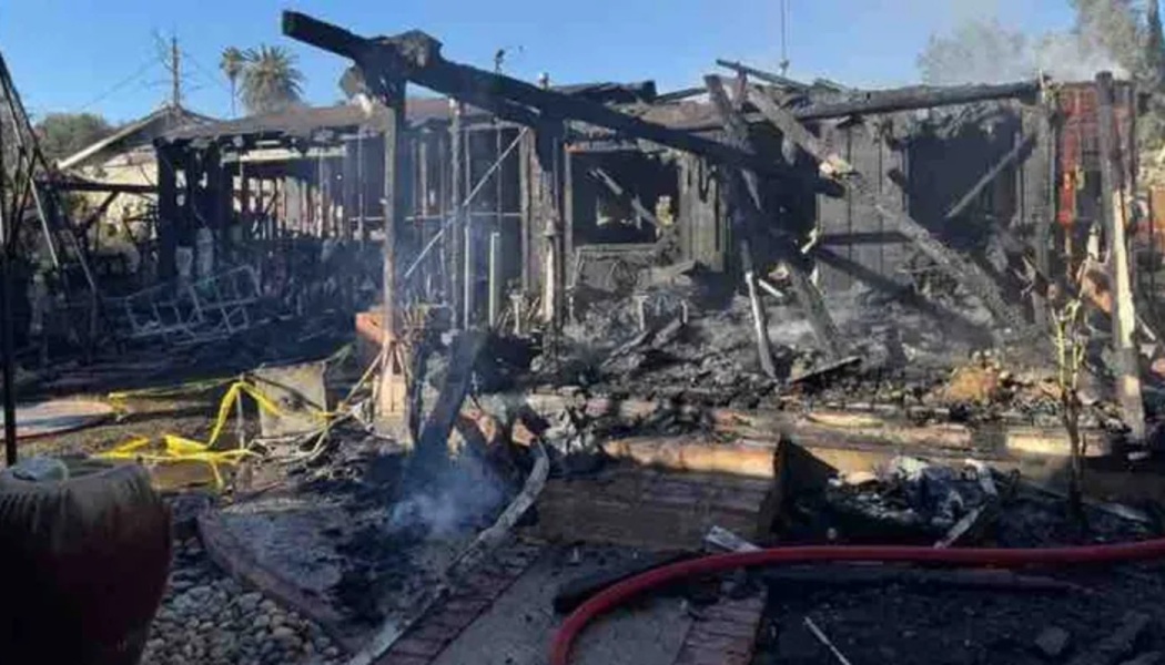 Charred, burnt remains of a house completely destroyed by a fire, with smoke still rising from the debris.
