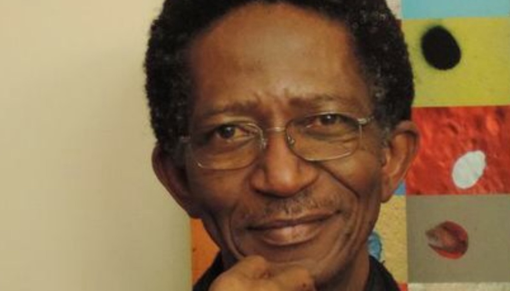Close-up portrait of a Black man with glasses smiling gently with his hand resting under his chin.