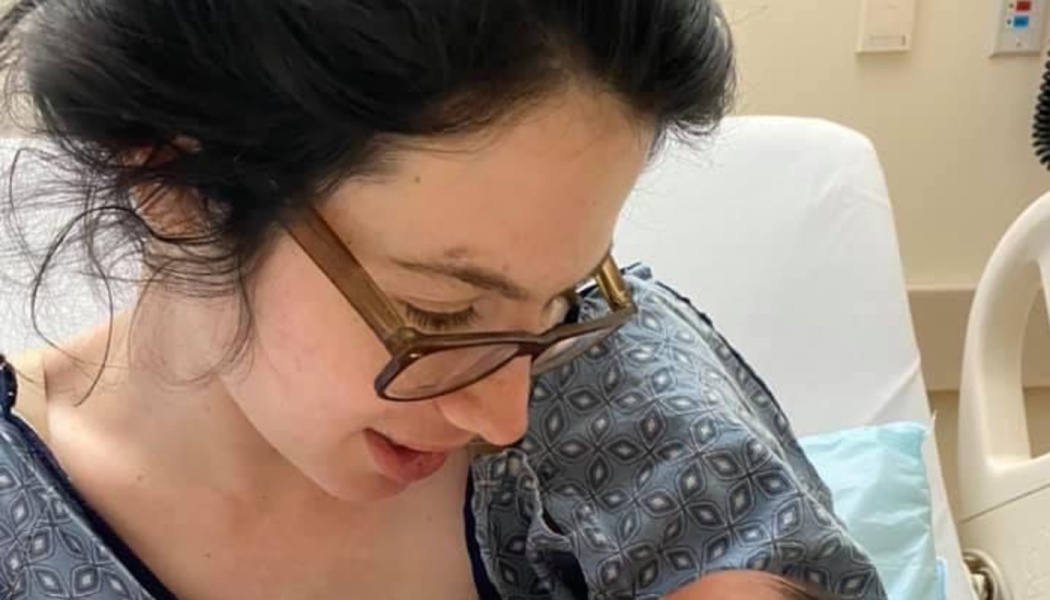 A dark-haired woman wearing glasses and a hospital gown looks down lovingly at her newborn baby.