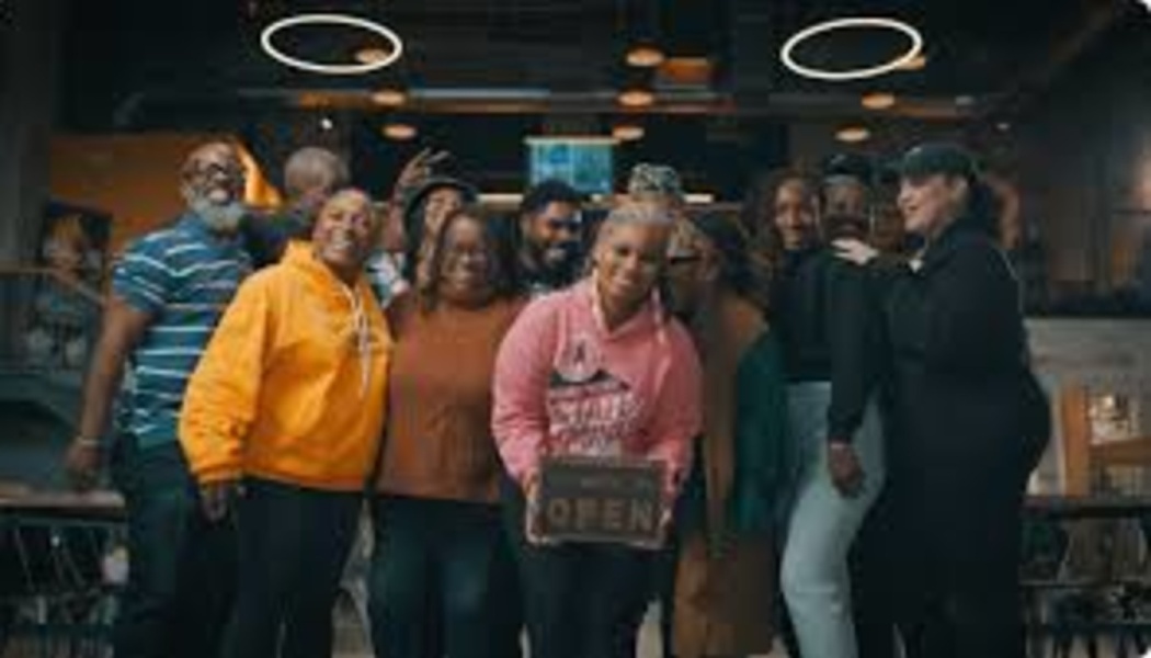 A diverse group of smiling people standing together indoors, with a woman in the center holding a small OPEN sign.