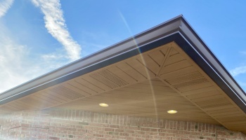 Brown soffit with recessed lighting and dark gutters on a brick exterior wall against a clear blue sky.