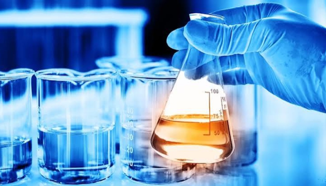 A gloved hand holds an Erlenmeyer flask filled with glowing amber liquid in a laboratory with glass beakers.