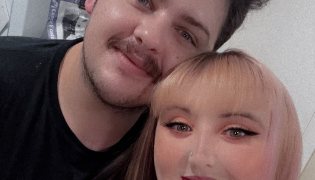 Close-up selfie of a smiling young couple, a man with a mustache and a woman with pinkish-blonde bangs.