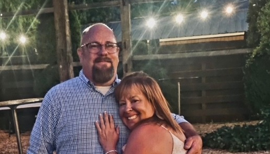 A smiling middle-aged couple embracing outdoors under glowing string lights.