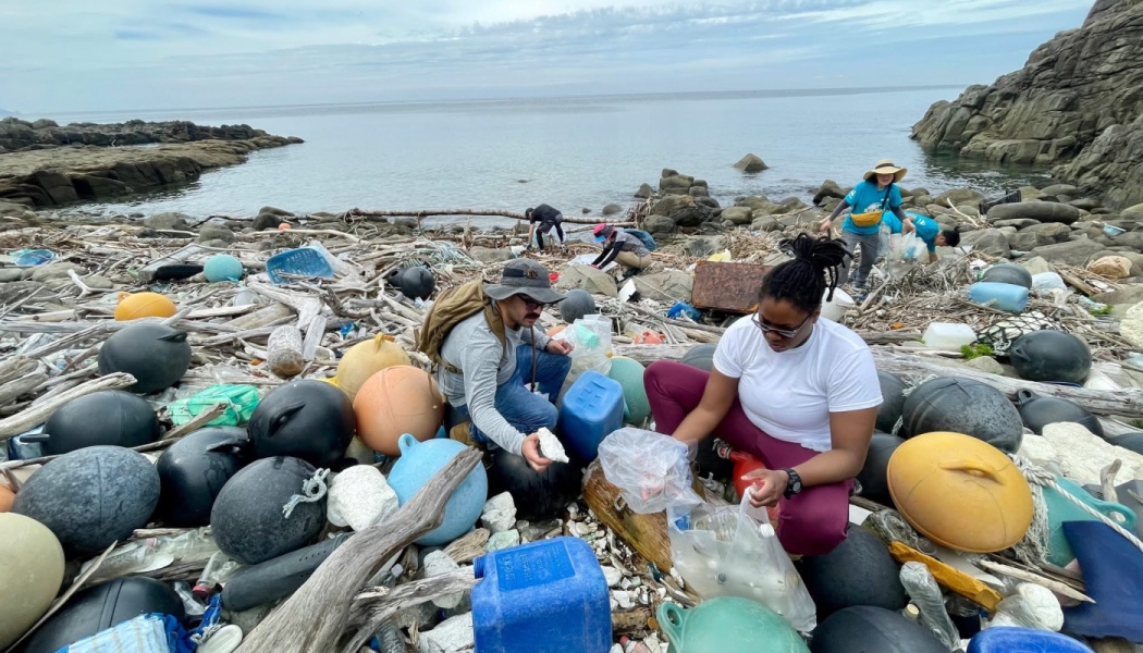 Volunteers pick up plastic trash, containers, and buoys from a littered rocky coastline.