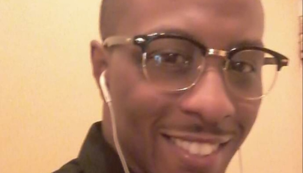 Close-up portrait of a smiling Black man wearing browline glasses and white earbuds.