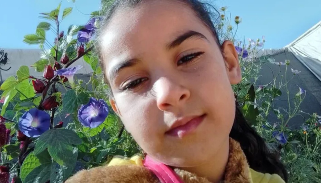 Close-up of a young girl with dark hair looking gently at the camera, surrounded by purple garden flowers.