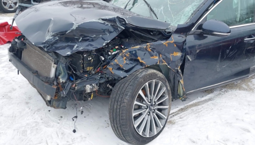 Severely damaged dark blue sedan with a smashed front end, crushed hood, and exposed engine on snow.