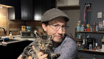 A smiling man wearing glasses and a flat cap holding a striped tabby cat in a kitchen.