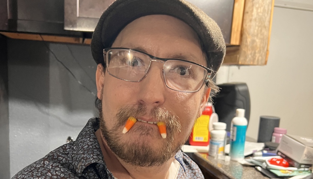 A bearded man in glasses and a flat cap with candy corn sticking out of his mouth like vampire fangs.