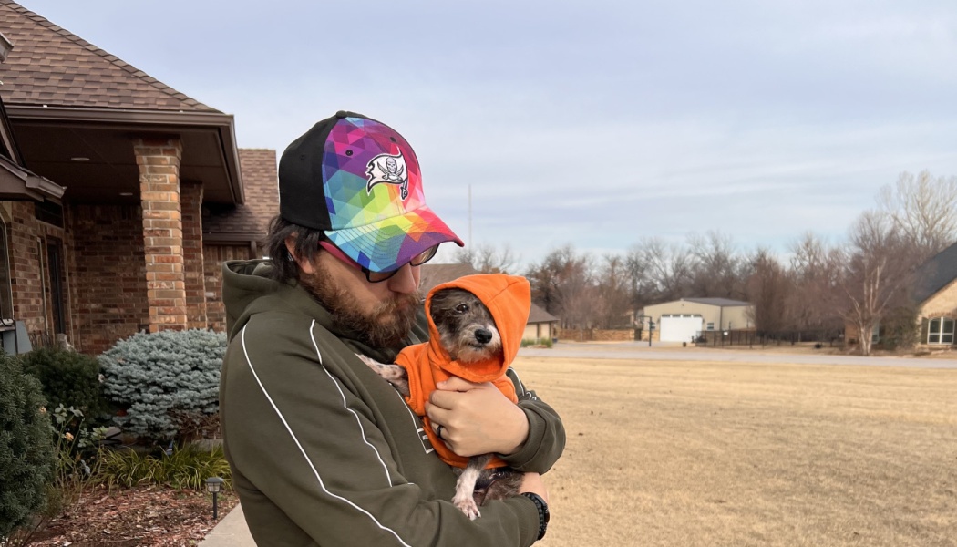 A bearded man wearing a colorful cap holds a small scruffy dog dressed in a bright orange hoodie outdoors.