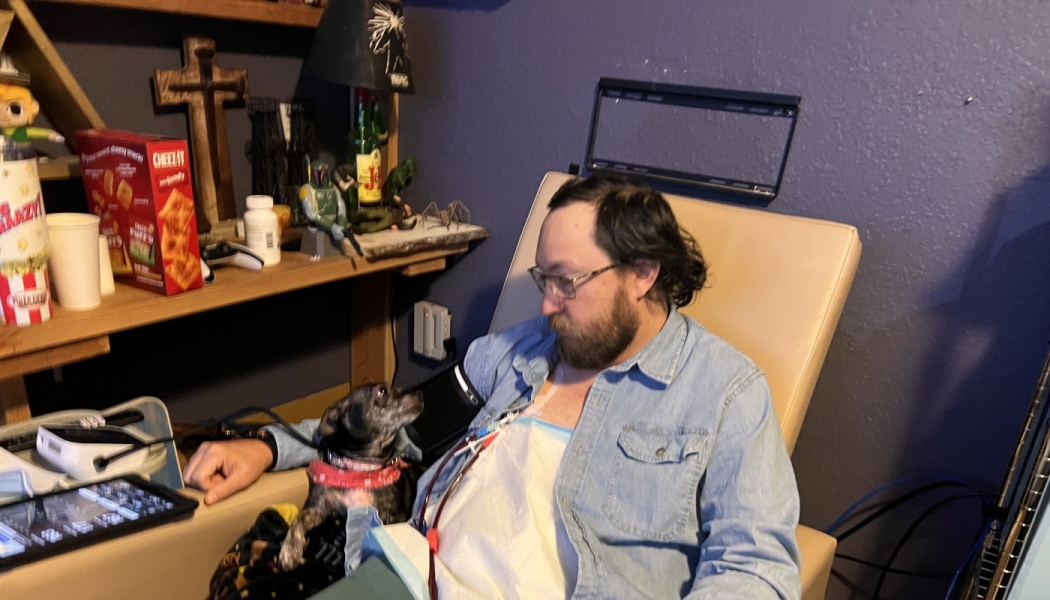 A bearded man receiving medical treatment in a chair looks affectionately at his small dog sitting on his lap.