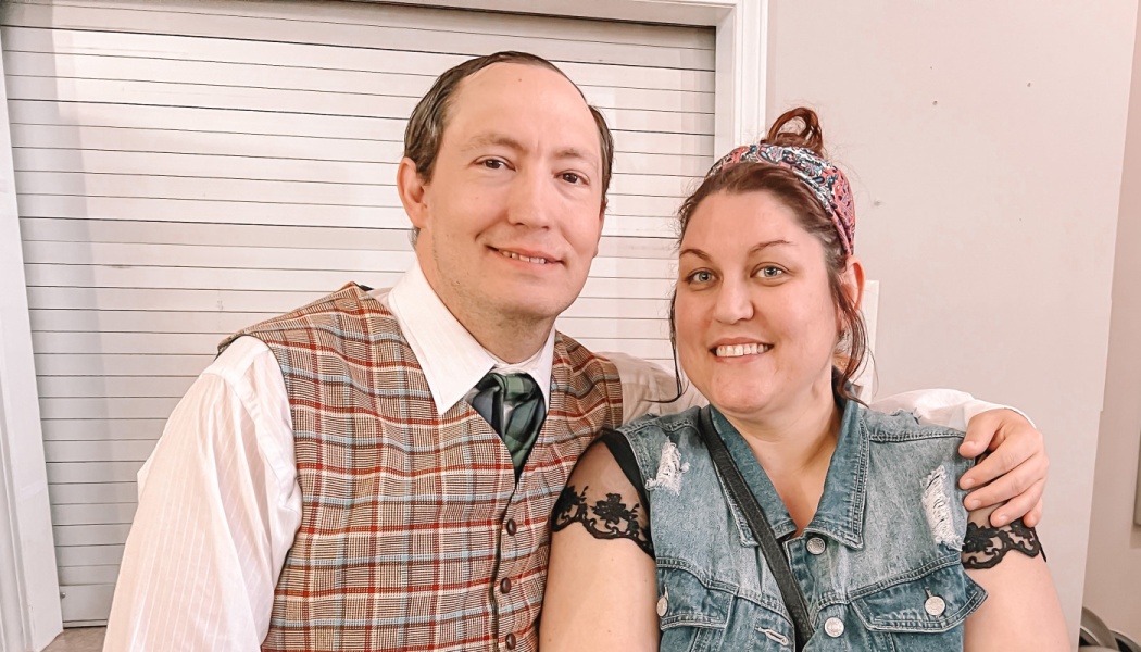 A smiling man in a plaid vest and tie poses with his arm around a smiling woman wearing a denim vest.