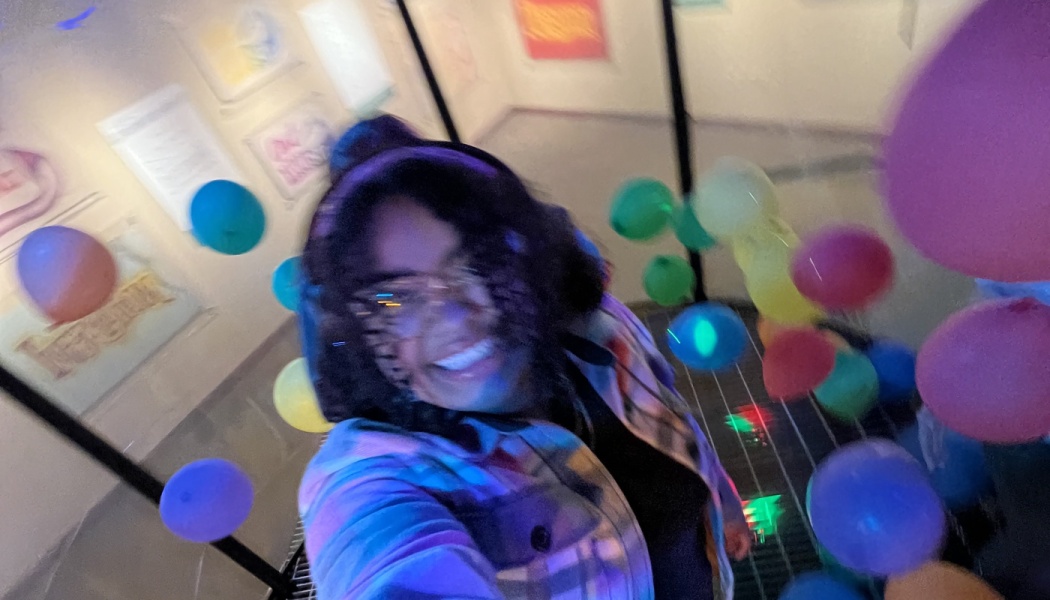 A smiling person wearing glasses takes a joyful selfie surrounded by bright, colorful floating balloons.
