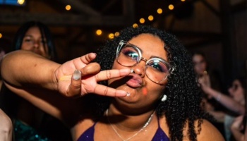 A young woman with curly hair and clear glasses throwing a peace sign and making a kissy face at a party.
