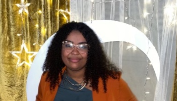 A smiling woman with dark curly hair and glasses in an orange blazer poses before a moon and star backdrop.