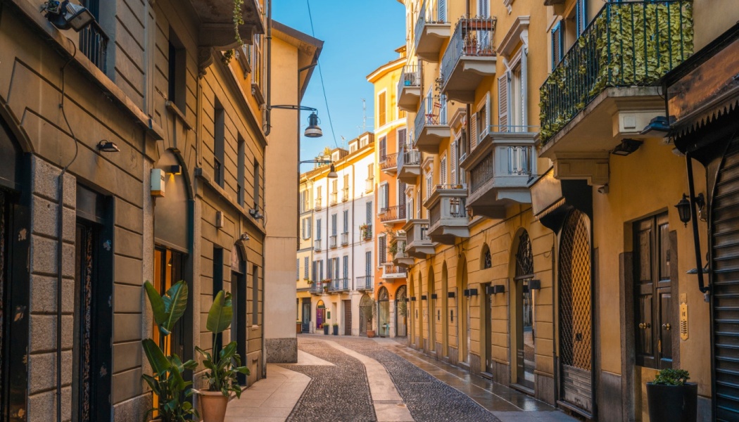 Narrow cobblestone street lined with warm yellow historic buildings, balconies, and potted green plants on a sunny day.