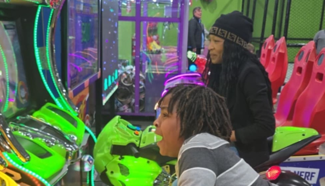 A young boy excitedly plays a green motorcycle arcade game beside a smiling woman.