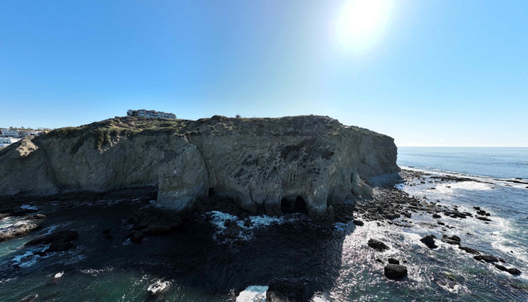 Sunlit rocky coastal cliffs with sea caves overlooking the ocean under a clear blue sky.