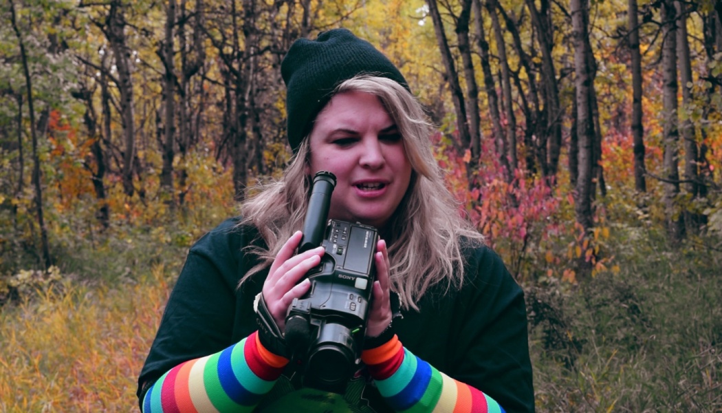 A person wearing a beanie and rainbow arm warmers holds a vintage camcorder in an autumn forest.