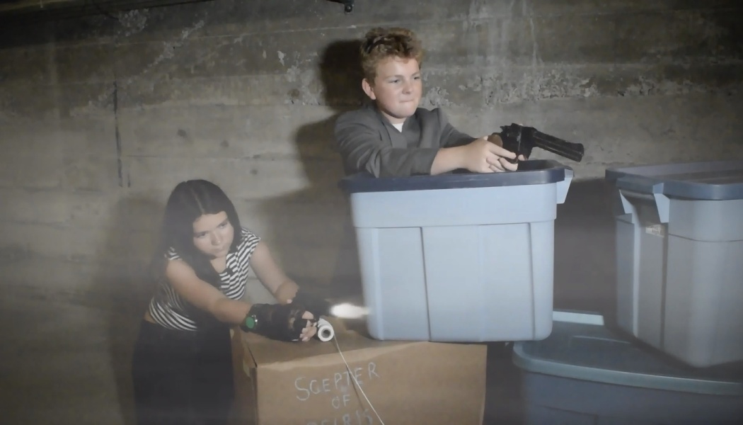Two children playing action heroes with toy guns behind storage bins in a foggy basement.