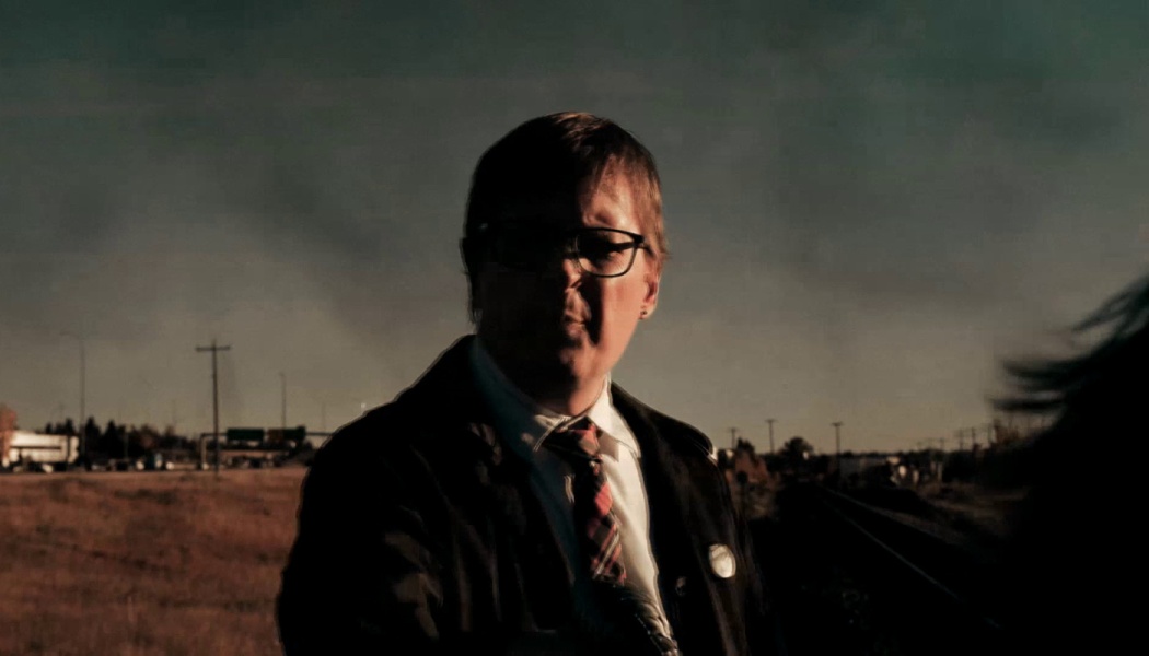 A person wearing glasses and a tie standing outdoors under a moody, shadowed sky at dusk.