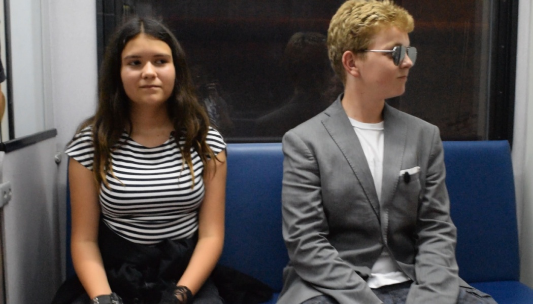 A girl in a striped shirt and a boy wearing a grey suit and sunglasses sitting on a train seat.