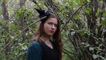 A young woman wearing a mini witch hat headband stands in a dense forest, looking off to the side.