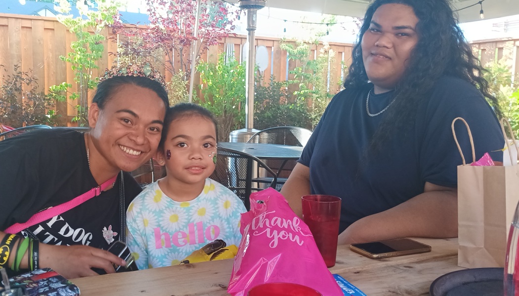 A smiling family of three, including a young child with face glitter, sitting together at an outdoor table.