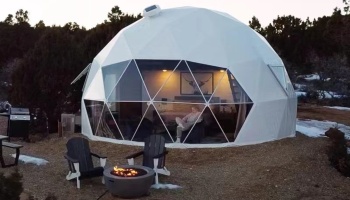 Cozy white geodesic glamping dome lit at dusk, featuring a warm interior, fire pit, and outdoor chairs.