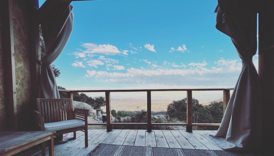 Wooden deck balcony with curtains and a bench, overlooking a wide valley view under a bright blue sky.