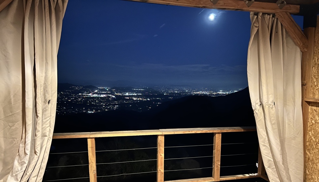 Nighttime view of glowing city lights and a bright moon framed by open curtains on a wooden balcony.
