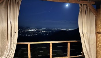 Nighttime view of glowing city lights and a bright moon framed by open curtains on a wooden balcony.