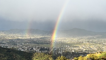 A vivid double rainbow stretches over a valley city framed by mountains under a cloudy sky.