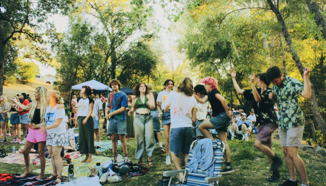 A group of young people joyfully dance together outdoors at a sunny, tree-lined park gathering.