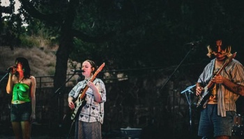 Three band members perform live outdoors at night, playing electric guitars and singing into microphones.