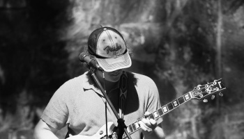 A musician wearing a Fender cap plays an electric guitar into a microphone in black and white.
