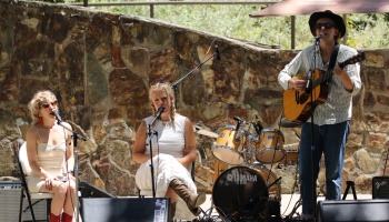 Three band members performing live outdoors with guitar, drums, and microphones in front of a stone wall.