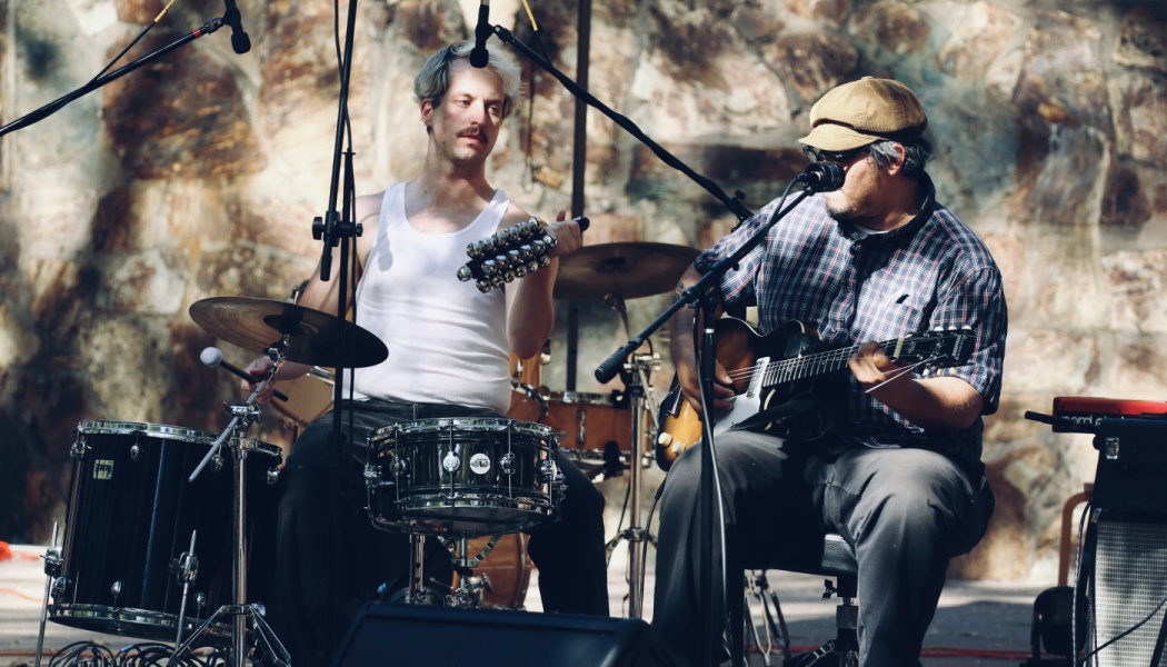 Two musicians perform outdoors: a drummer shaking sleigh bells and a seated guitarist singing into a microphone.