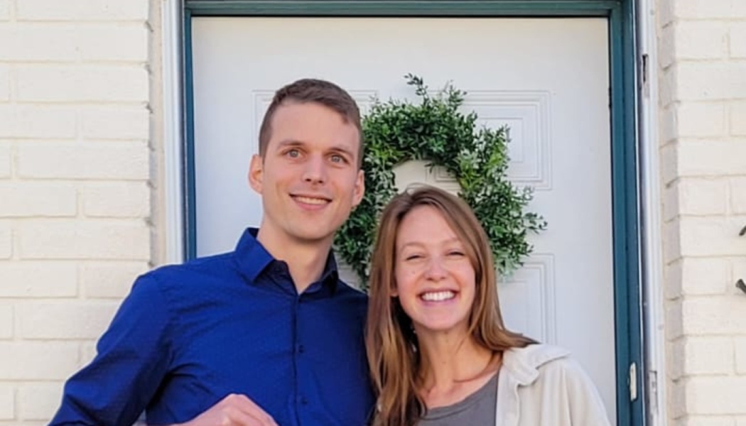 A smiling young couple standing together in front of a white front door adorned with a green wreath.