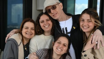 A man in a white cap and glasses stands with four smiling women gathered closely together in a group hug.
