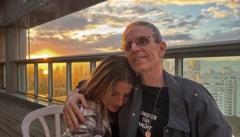 A man with glasses hugs a young woman resting her head against him on a city balcony at sunset.