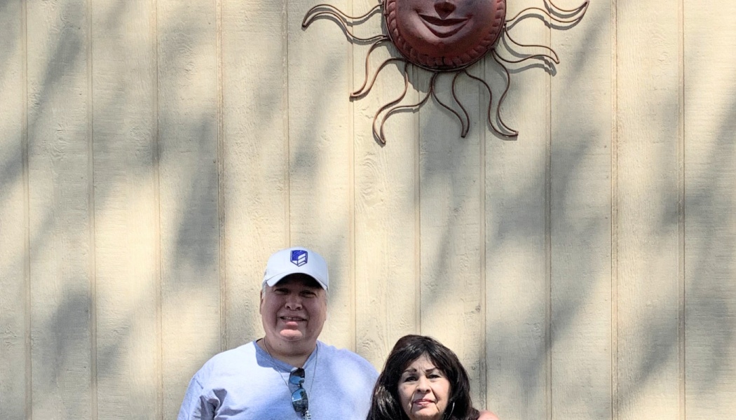 A smiling man and woman pose in front of a wooden wall with a decorative metal sun mounted above them.