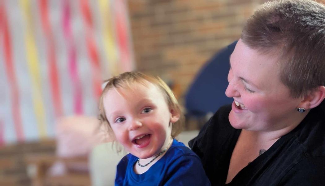 A happy toddler in a blue shirt smiles brightly as a smiling adult with short hair looks on joyfully.
